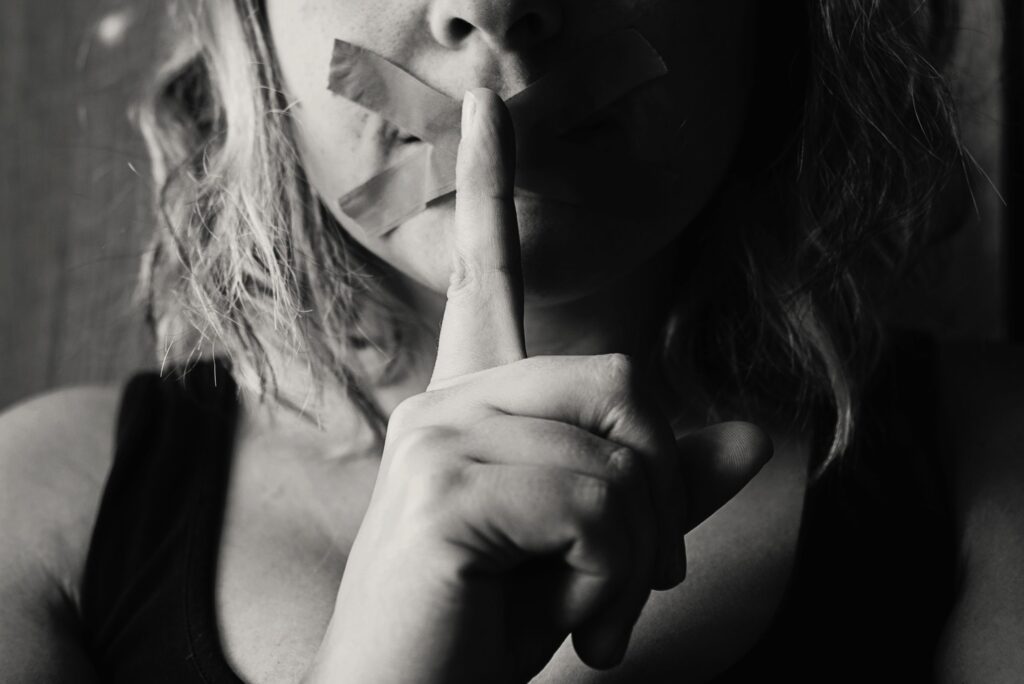 Black and white image of woman making the 'shhh' sign with tape over mouth